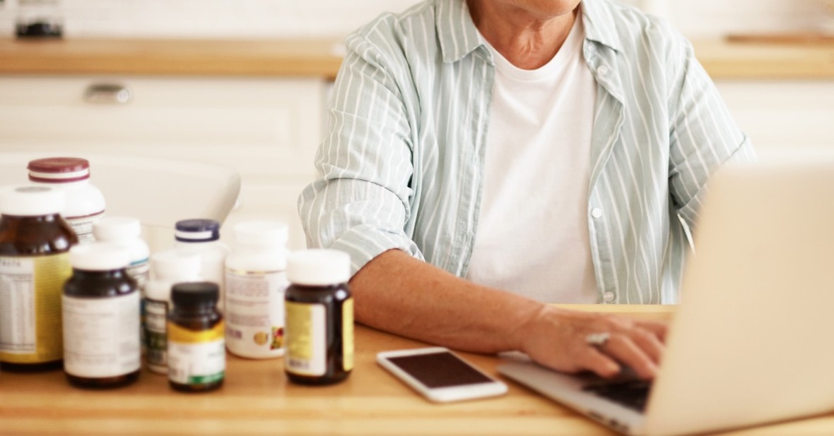 Persona mayor utilizando un ordenador al lado de varios botes de suplementos de vitaminas, para combatir una posible desnutrición por la interacción entre medicamentos y alimentación
