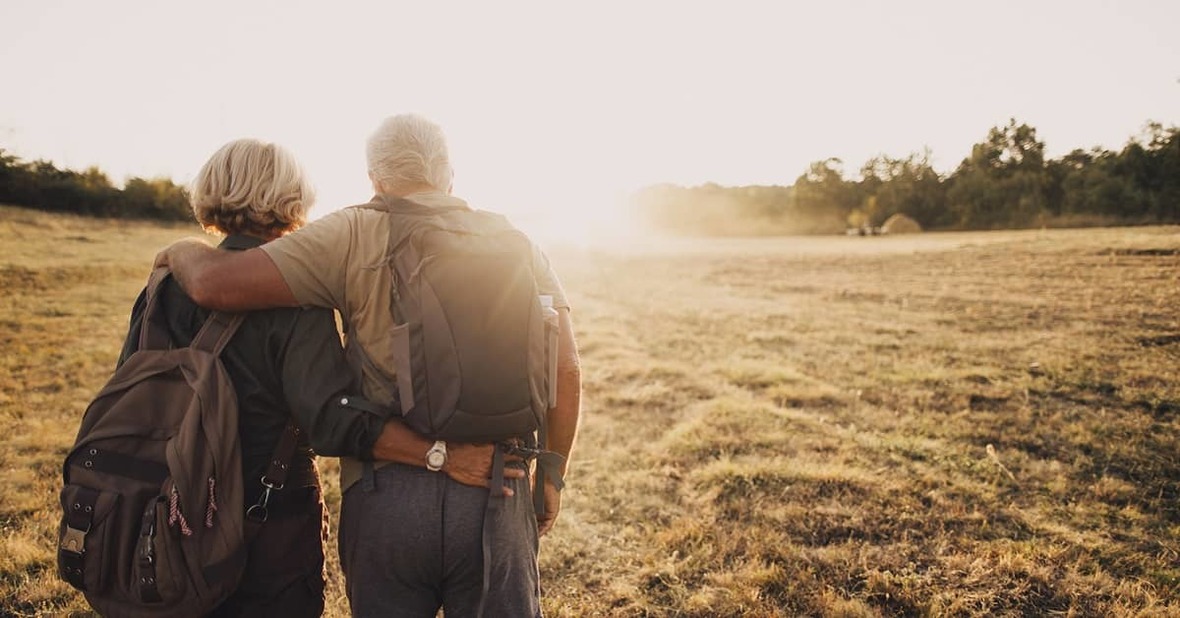 Pareja de personas mayores caminando por el campo abrazados con mochilas y ropa de senderismo
