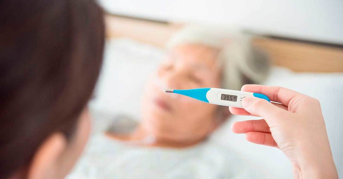 Cuidadora tomándole la temperatura a una persona mayor con fiebre tumbada en la cama