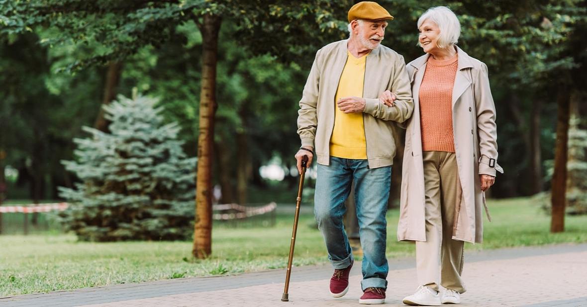 Pareja de personas mayores paseando por el parque agarrados del brazo porque el hombre sufre un problema de movilidad y lleva un bastón