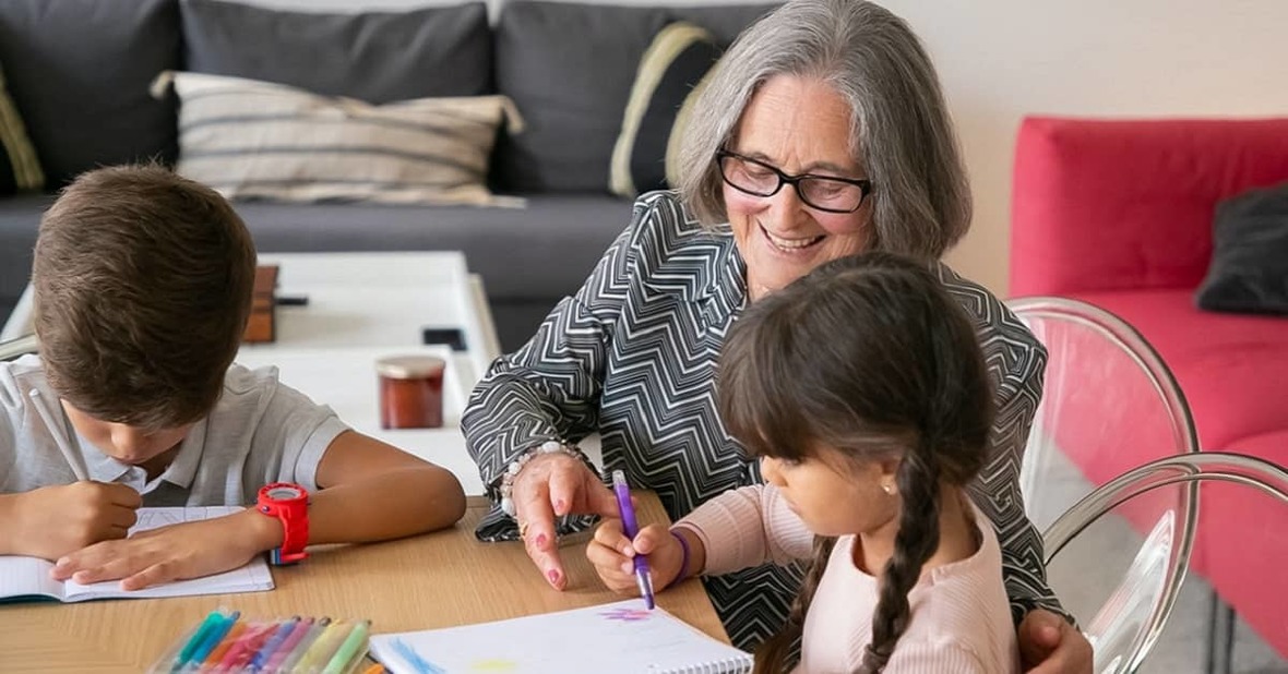 Abuela ayudando a sus dos nietos a dibujar, sentados en la mesa del salón